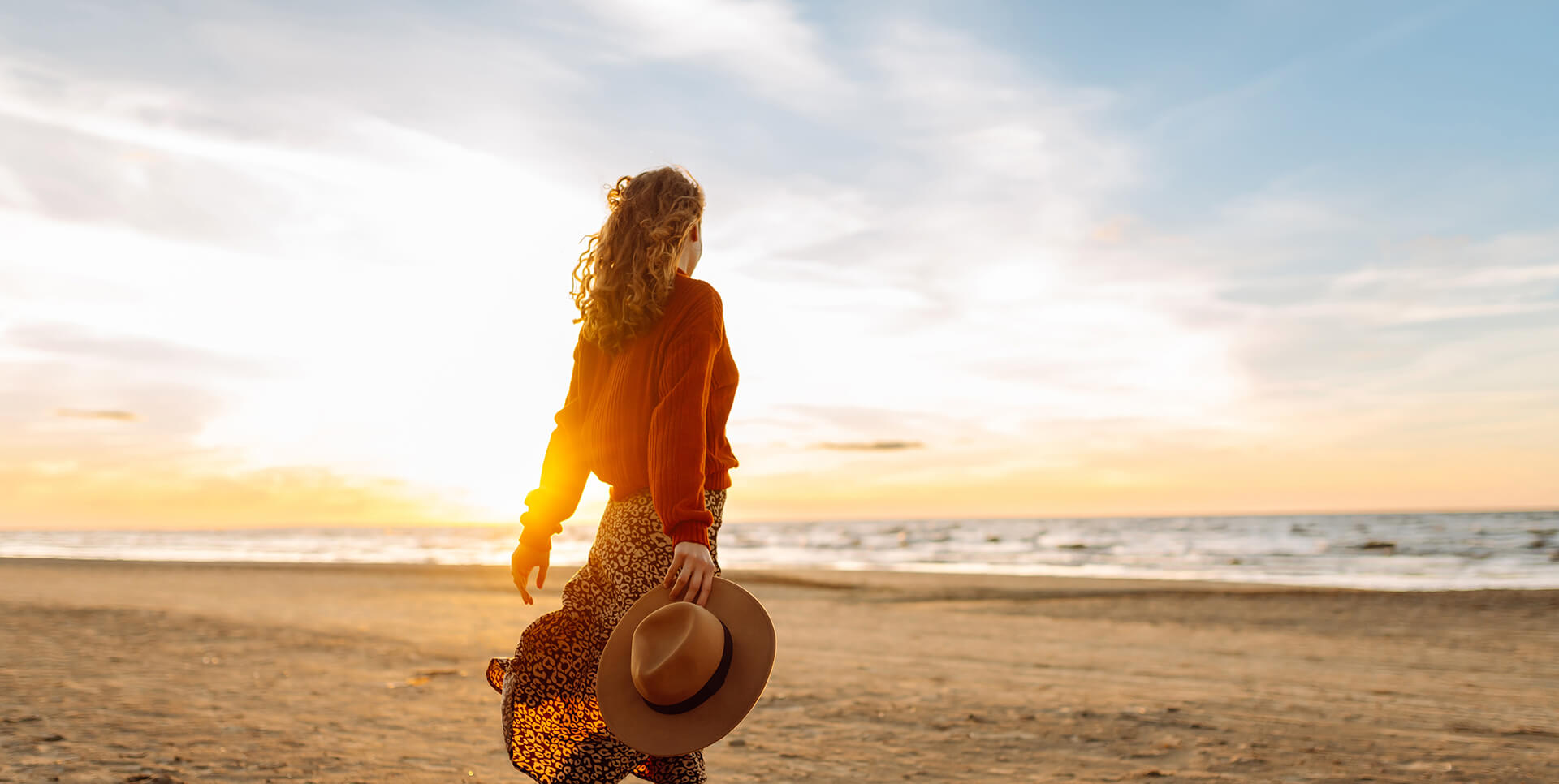 Belha Collection - Femme marche sur la plage un chapeau à la main, face au coucher de soleil